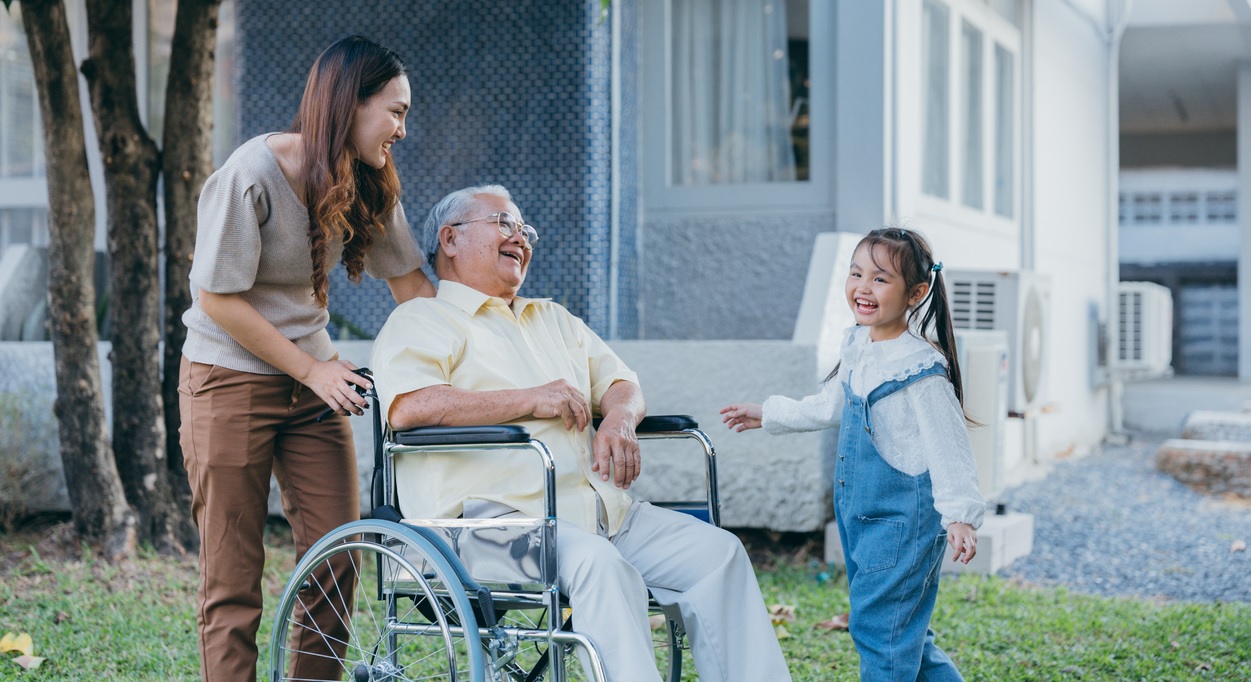 Asian woman pushing older man in wheelchair with younger asian girl smiling next to man