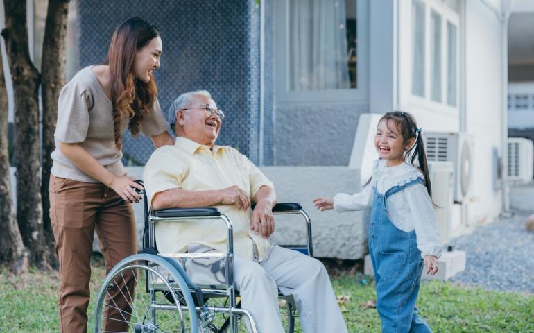 Asian woman pushing older man in wheelchair with younger asian girl smiling next to man